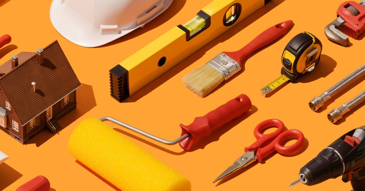 Construction tools like a level, a paintbrush, scissors, a paint roller and more, laid on a bright orange text